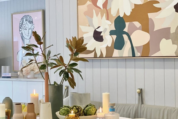 Modern dining room table styled with assorted colored glass vases, candles, and decorative items under floral wall art in soft neutral tones, showcasing the colour of home collection.