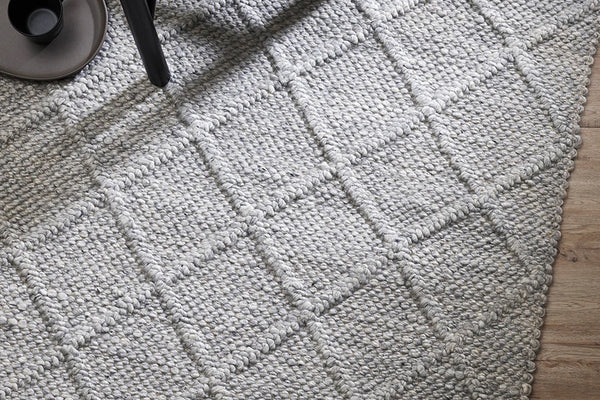 Textured gray woven area rug on wooden floor with black chair leg and tray nearby, representing Baya working with Good Weave commitment
