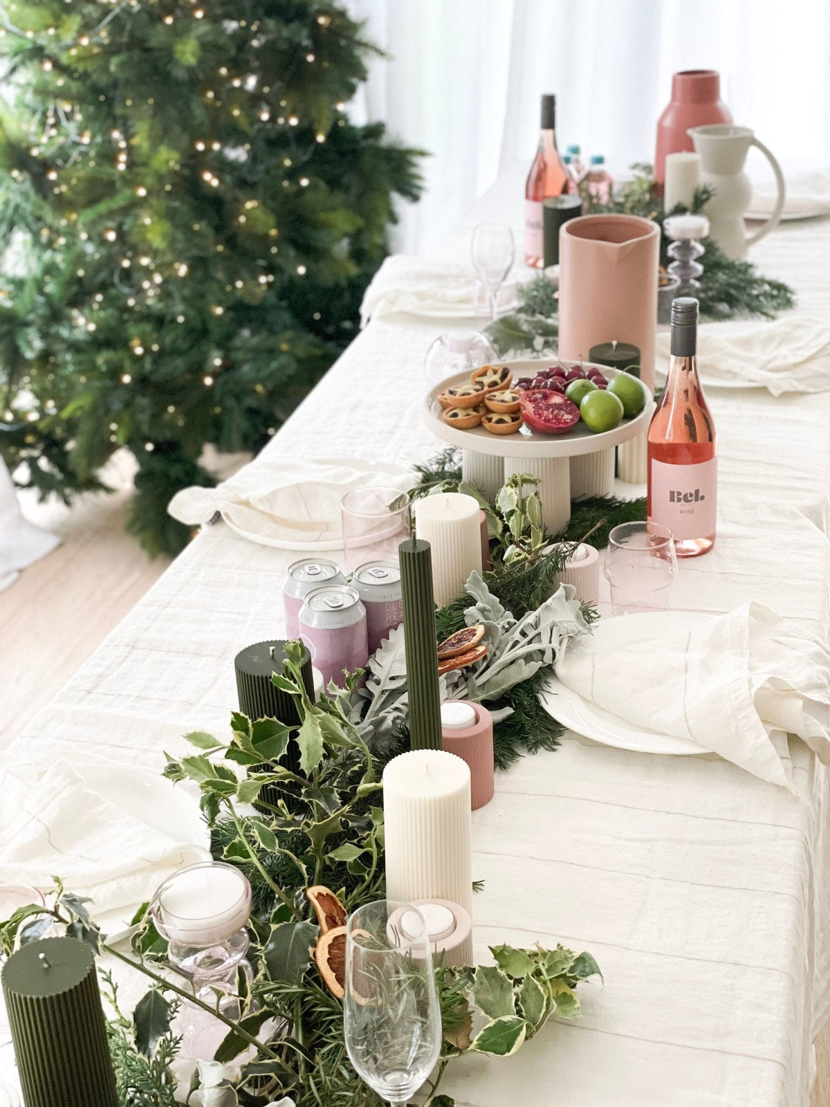 Festive holiday table setting with blush linen, greenery, candles, and rosé wine by a decorated Christmas tree