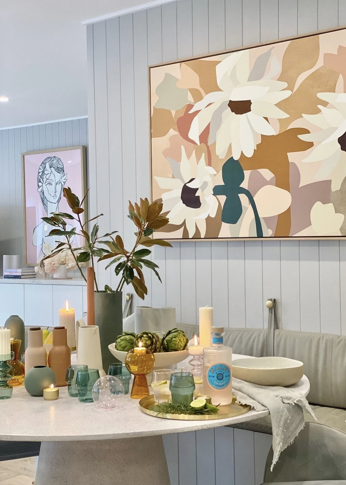 Modern dining room table styled with assorted colored glass vases, candles, and decorative items under floral wall art in soft neutral tones, showcasing the colour of home collection.