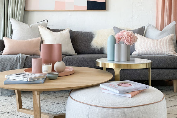 Modern living room with a round beige ottoman, wooden coffee table, gray sofa, and pastel decor accents
