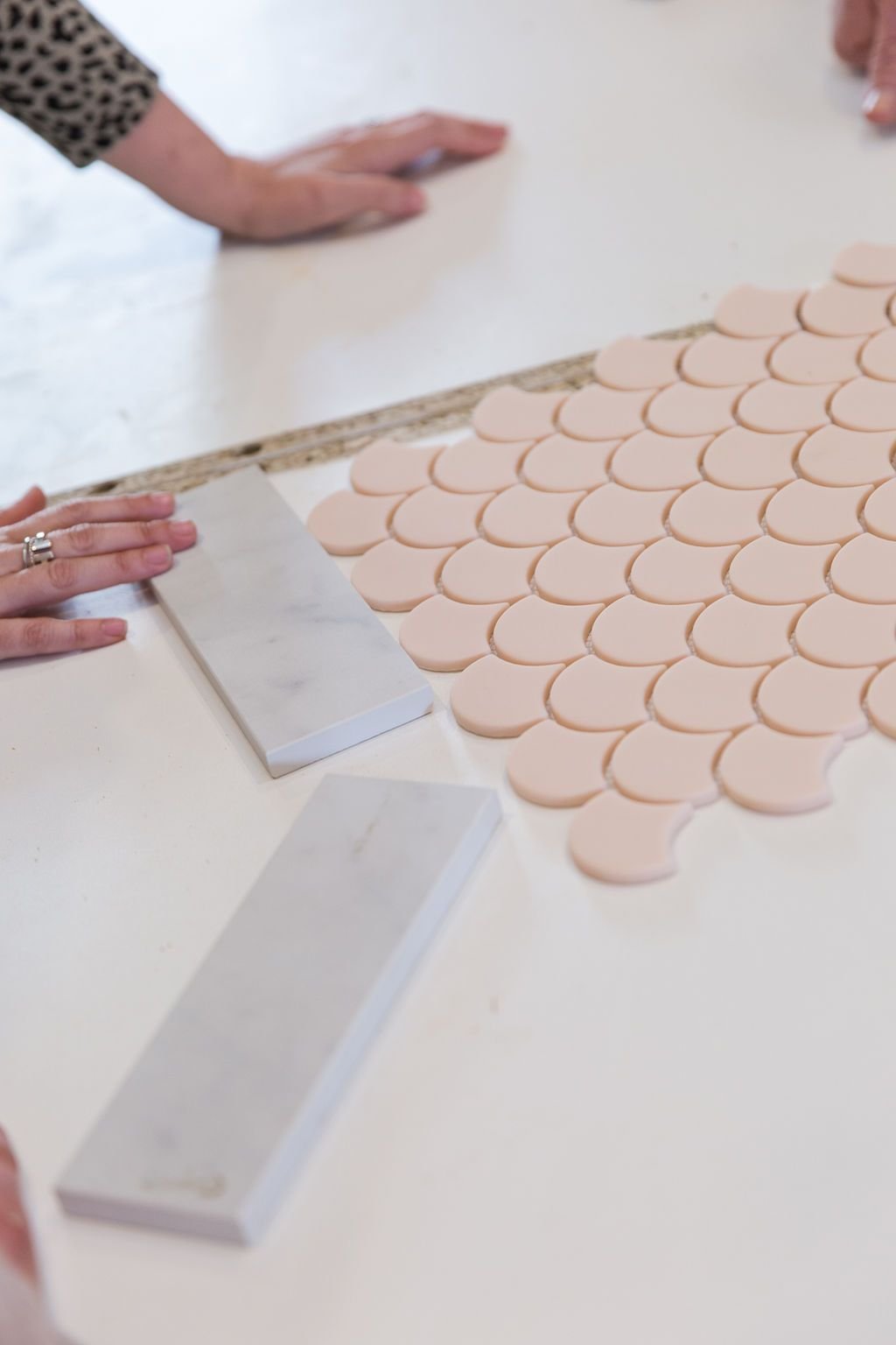 Hands arranging blush pink fish scale mosaic tiles and marble samples on a white surface for kitchen renovation Project Seven Update