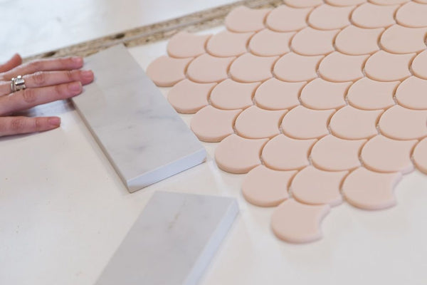 Hands arranging blush pink fish scale mosaic tiles and marble samples on a white surface for kitchen renovation Project Seven Update