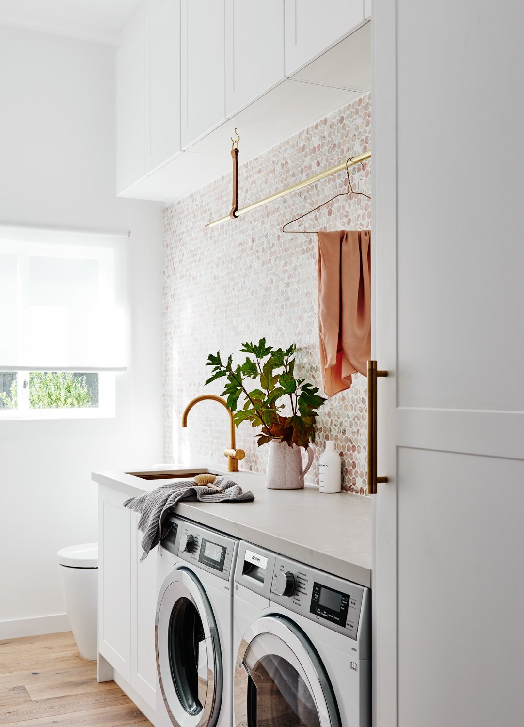 Modern laundry room with white cabinetry, front-loading washer and dryer, gold faucet, and decorative backsplash featuring a plant on the countertop norsuHOME