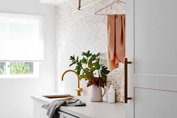 Modern laundry room with white cabinetry, front-loading washer and dryer, gold faucet, and decorative backsplash featuring a plant on the countertop norsuHOME