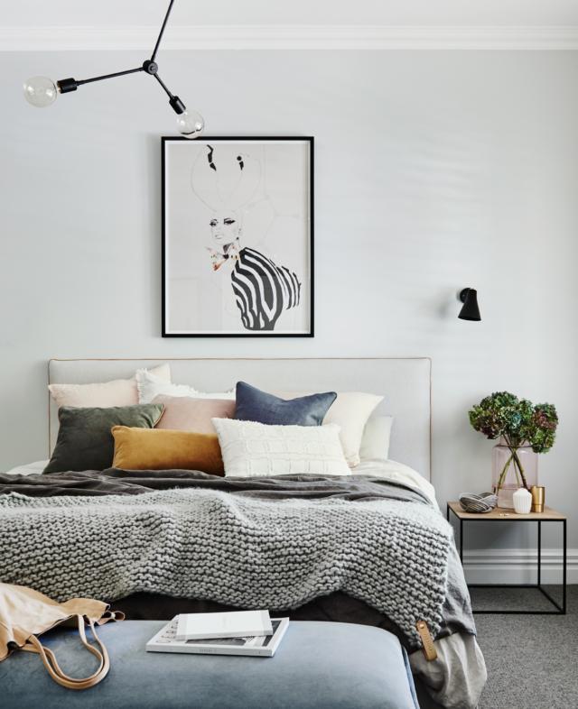 Modern minimalist main bedroom with grey knit blanket, assorted pillows, framed abstract artwork, black wall sconces, and bedside table with plant, showcasing norsuHOME design.