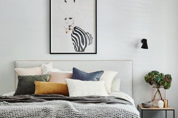 Modern minimalist main bedroom with grey knit blanket, assorted pillows, framed abstract artwork, black wall sconces, and bedside table with plant, showcasing norsuHOME design.