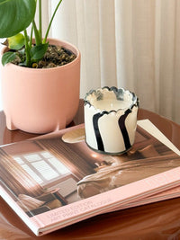 Ella Glass Candle in ivory and black with a sculptural hand-painted striped glass jar on a table next to a plant and book