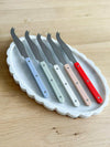 Elke Marble Tray oval white marble tray with scalloped edges holding five cheese knives with colorful handles on wood surface