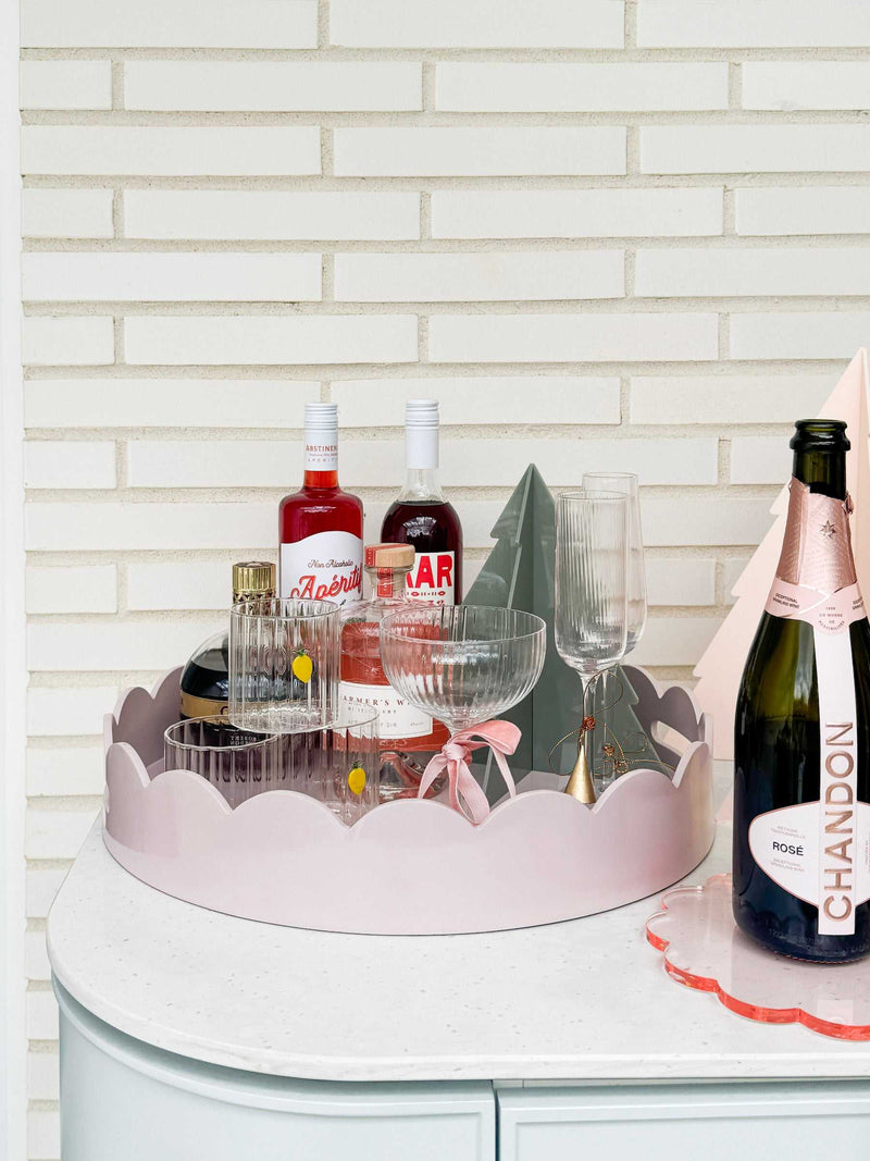 norsu Studio Round Scalloped Tray in Soft Violet with liquor bottles and glassware on a white surface against a brick wall background