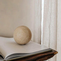 Beige travertine Orbit Sphere Table Sculpture on an open book with soft natural light background