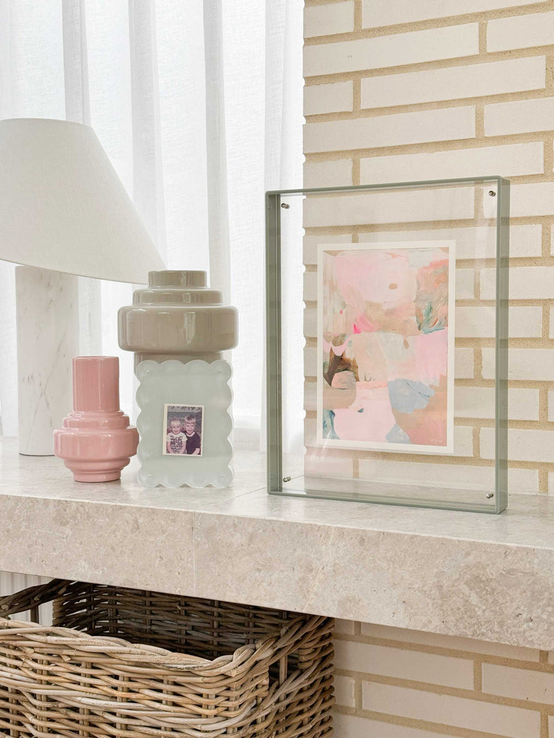 norsu STUDIO sage acrylic box gallery frame displaying pastel artwork on a mantel with decorative vases and wicker basket below