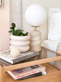 Orbit Sphere Table Sculpture in beige travertine displayed on a glass table next to a white textured planter and books.