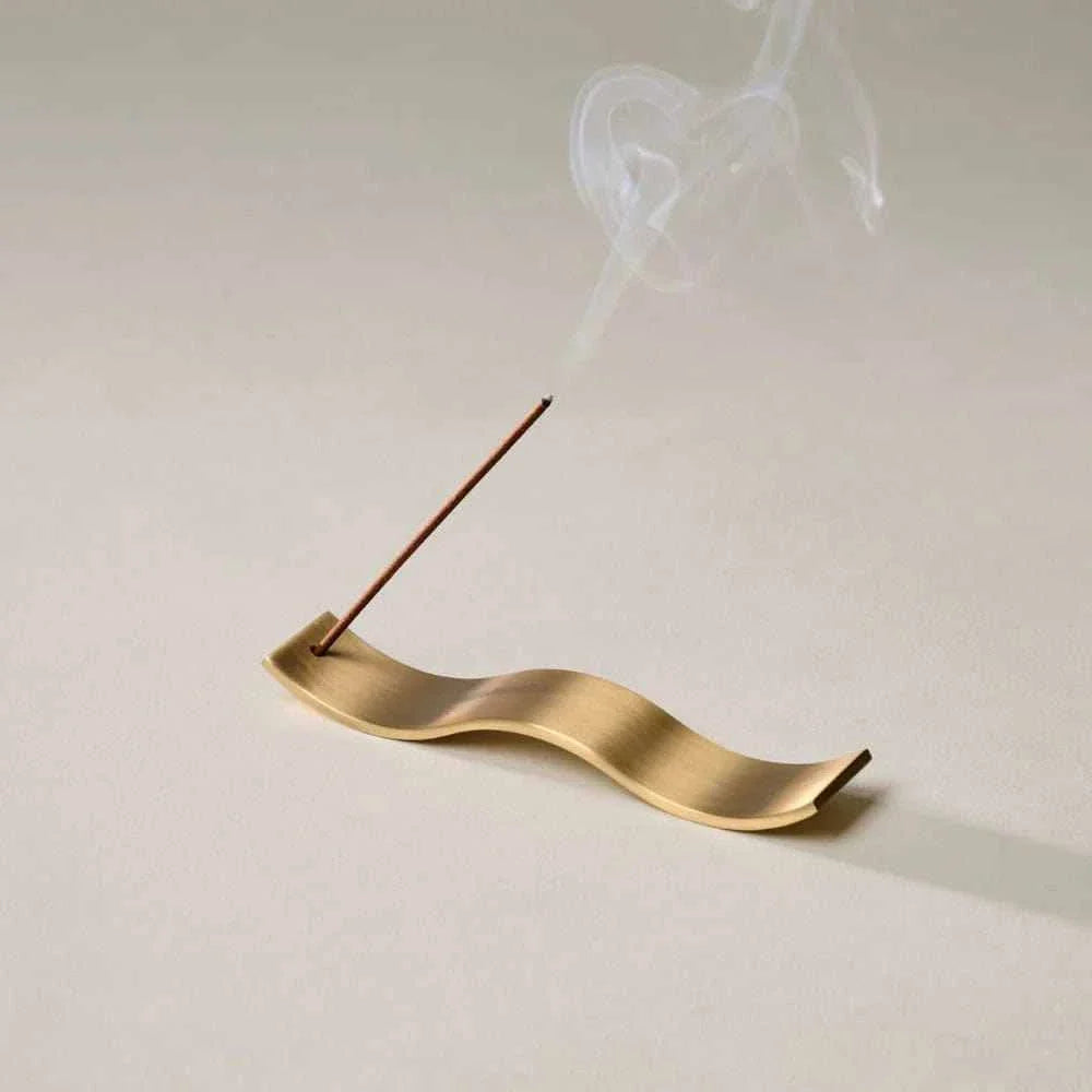 Gentle Habits Brass Wave Incense Holder in smooth wavy brass design holding a lit incense stick with smoke on a neutral background