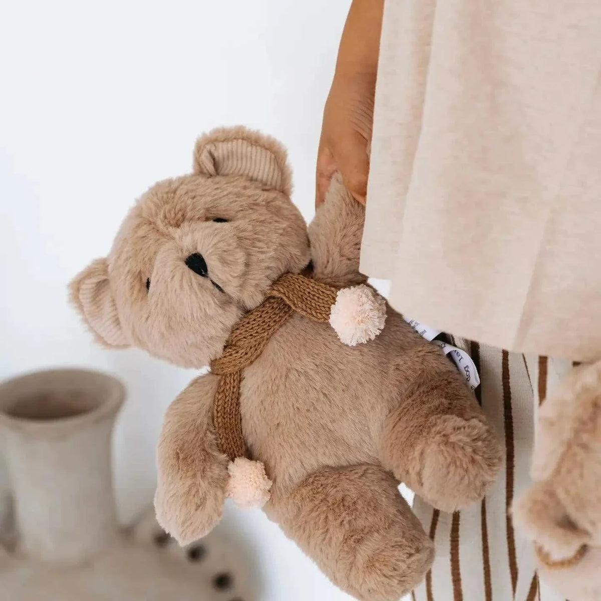Soft caramel plush Prudence Bear wearing a knitted chocolate merino wool scarf with pink pom poms held by a child