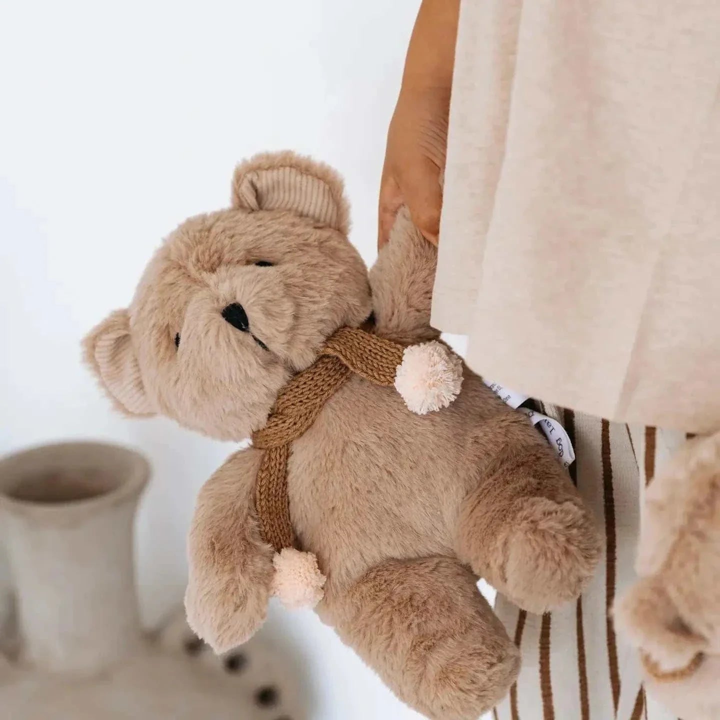 Soft caramel plush Prudence Bear wearing a knitted chocolate merino wool scarf with pink pom poms held by a child