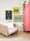 Modern living room with customizable wall art prints featuring a framed number 20 and "One Shot" text design next to a pink locker and beige sofa