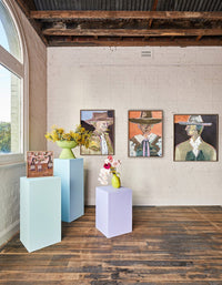 Gallery display of Marcia Priestley Limited Edition fine art canvas prints featuring three portraits with floating oak frames and decorative floral arrangements on pastel pedestals