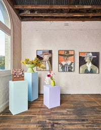 Marcia Priestley limited edition fine art canvas prints displayed on gallery wall with floral arrangements on pastel pedestals in rustic room