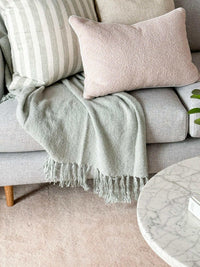 norsu Studio Cushion in blush boucle with white leather piping on a grey sofa with matching striped cushion and green throw blanket