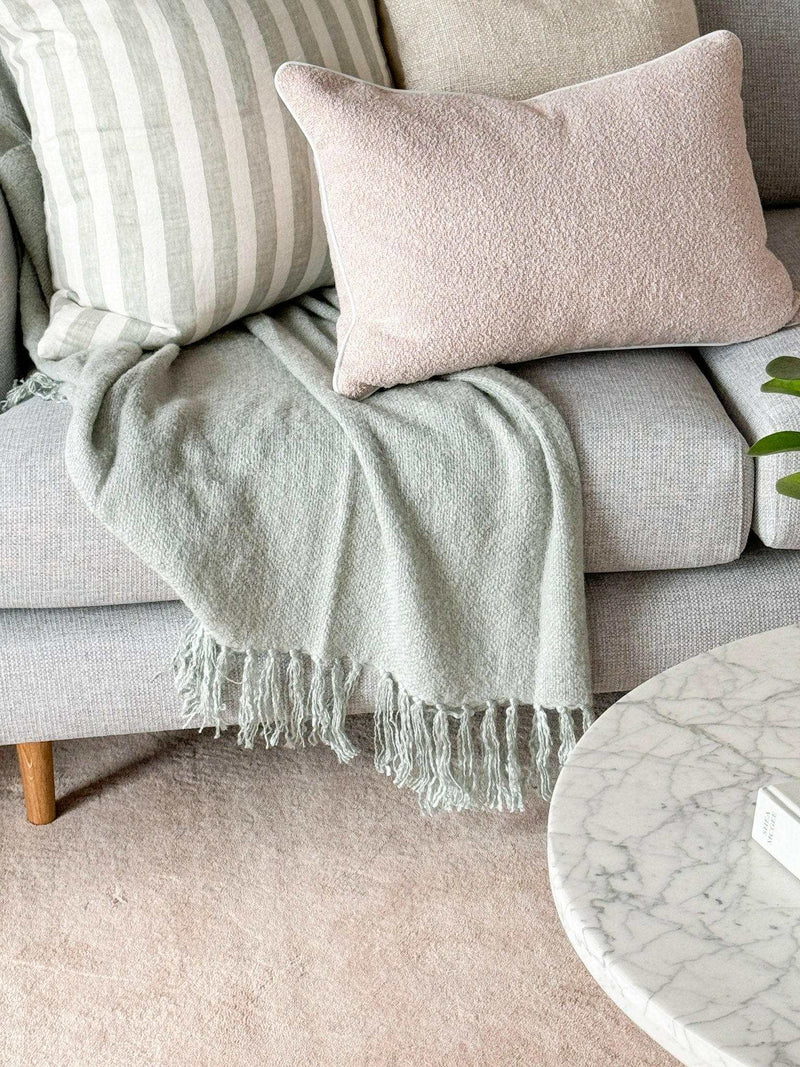 norsu Studio Cushion in blush boucle with white leather piping on a grey sofa with matching striped cushion and green throw blanket