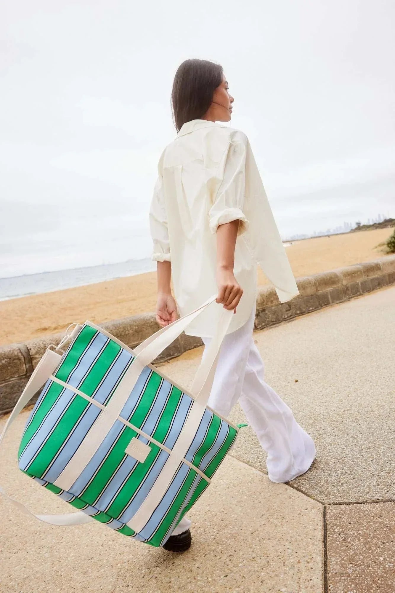 Base Supply Cooler Base Tropea Stripe insulated tote bag with green and blue stripes carried by woman at beach pathway