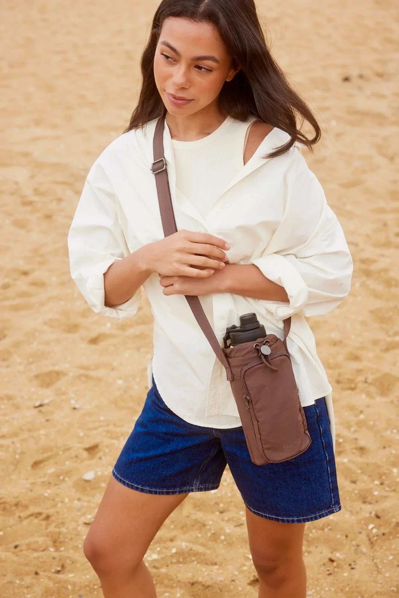 Woman wearing Base Supply Bottle Sling crossbody bag in chocolate color on beach carrying water bottle and essentials
