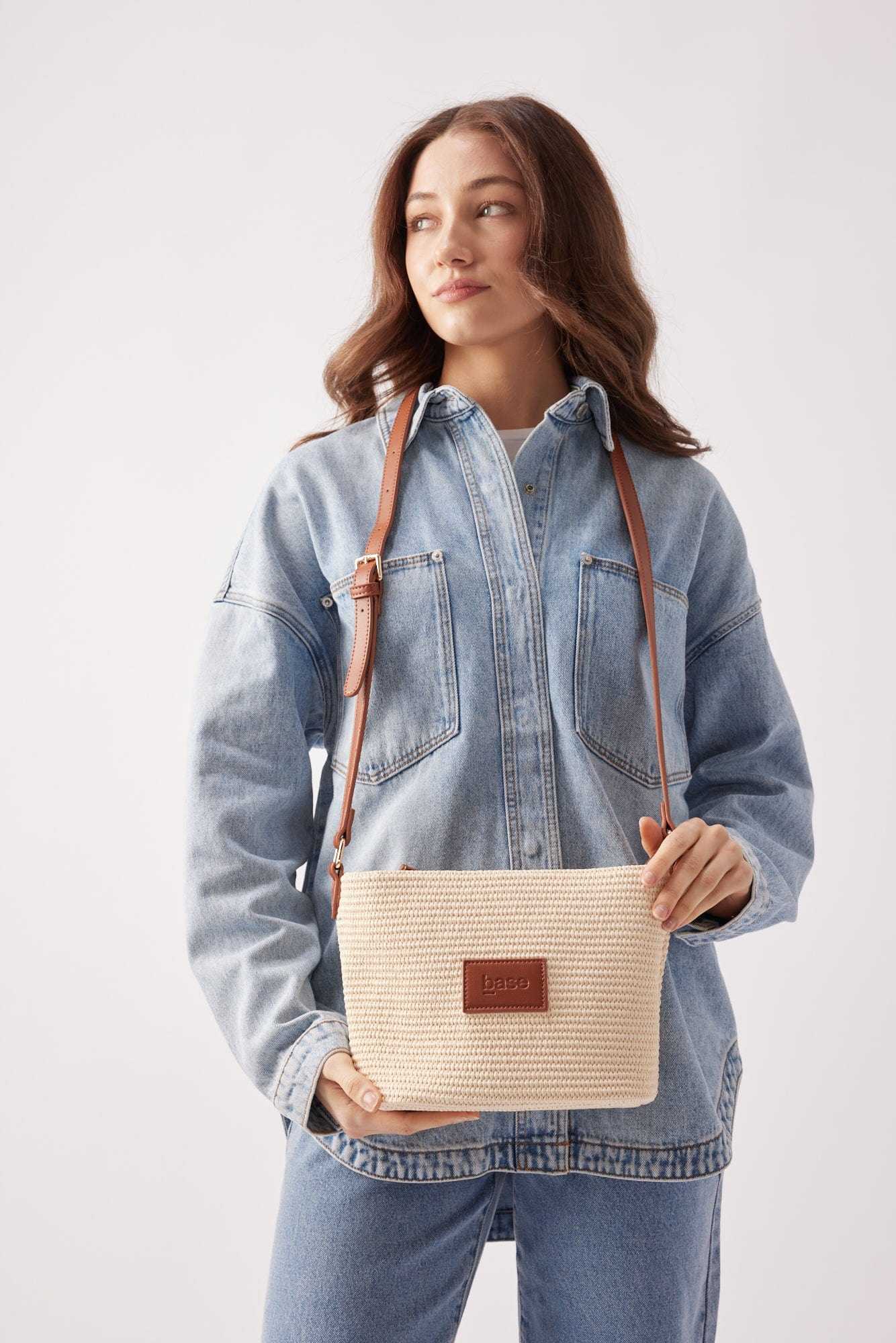 Woman wearing denim jacket holding tan Base Supply Panier Crossbody bag with woven straw texture and adjustable leather strap