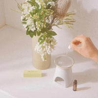 Gentle Habits diffuser oils being added to a ceramic aromatherapy oil burner near a vase of white flowers and a small oil bottle on a white countertop