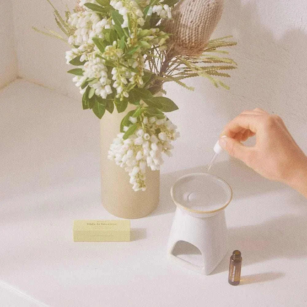 Gentle Habits diffuser oils being added to a ceramic aromatherapy oil burner near a vase of white flowers and a small oil bottle on a white countertop
