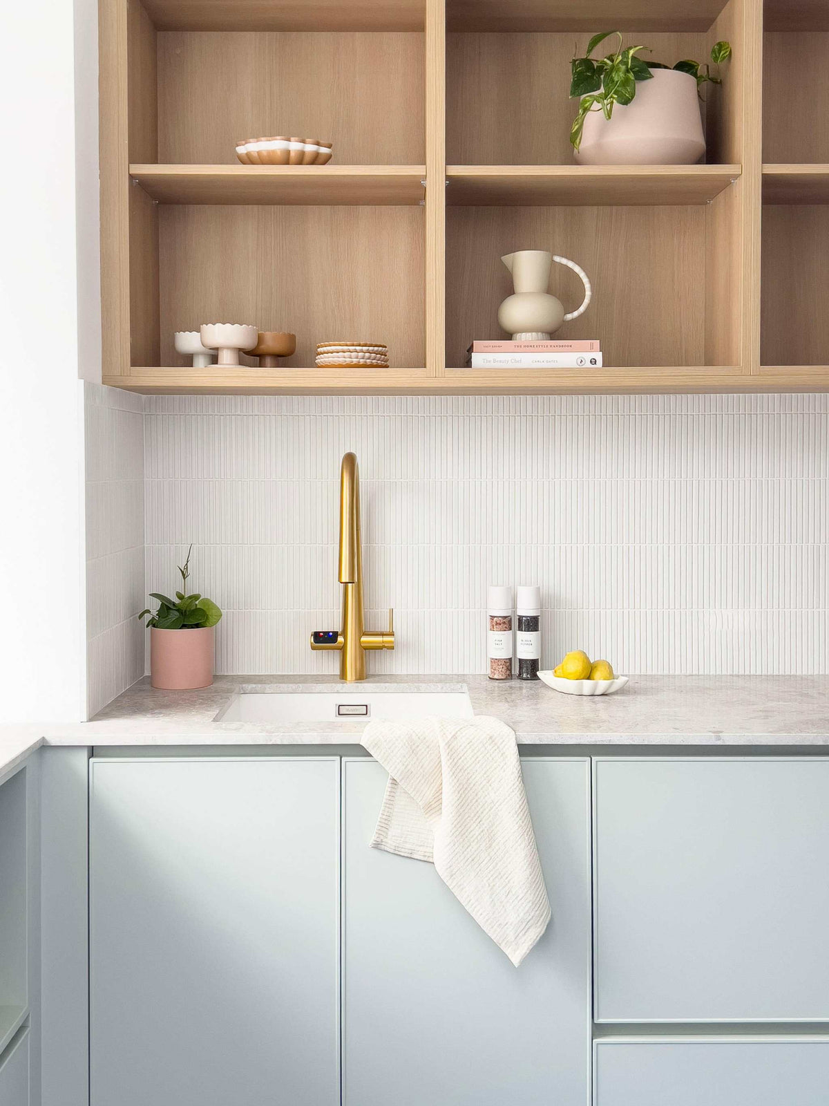 Minimalist kitchen shelves styled with neutral ceramics and green plants above a modern countertop with a gold faucet.