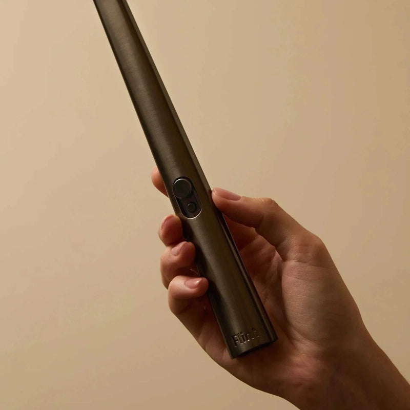Hand holding a sleek gunmetal Flint rechargeable lighter with a button and LED indicator, designed for lighting candles, paper, incense, and stoves.