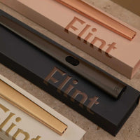 Sleek gunmetal Flint rechargeable lighter on black branded box with nearby pink and gold Flint lighter boxes.