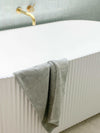 Paros Bath Mat in limestone color hanging on white modern bathtub with gold faucet