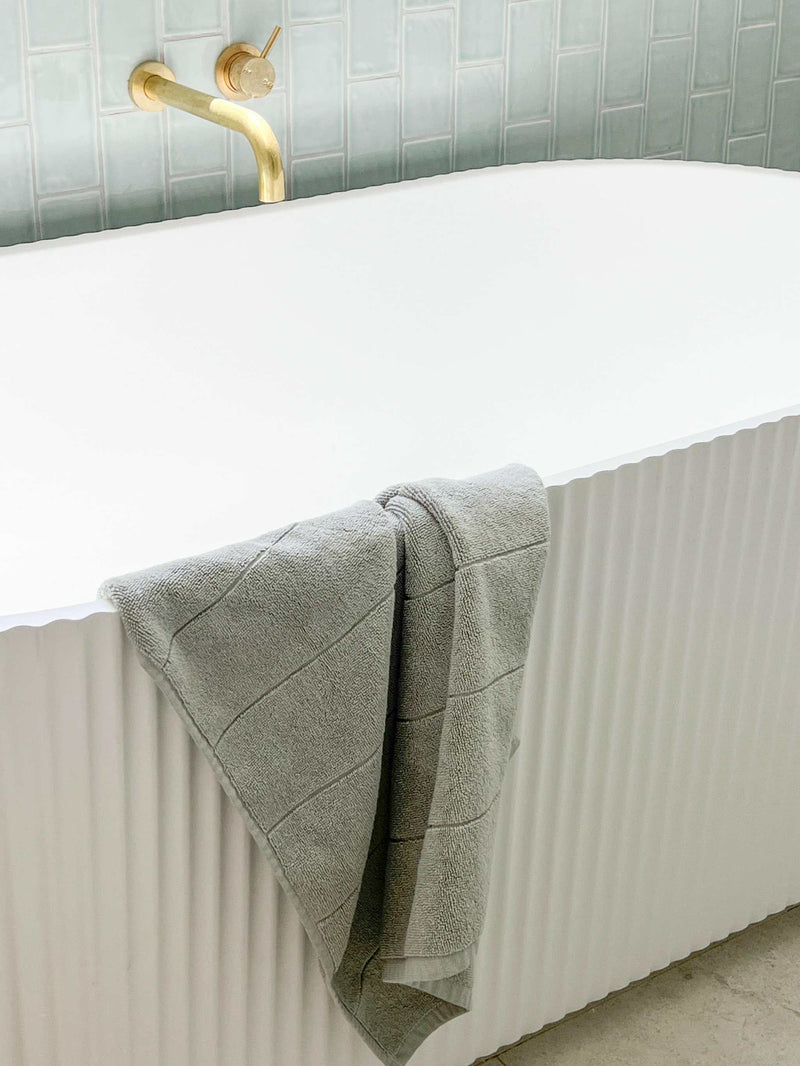 Paros Bath Mat in limestone color hanging on white modern bathtub with gold faucet
