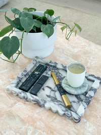 Large Palazzo scalloped marble bar tray in Viola marble with remote controls, lighter, and a candle on a coaster next to a potted green plant on a tiled surface