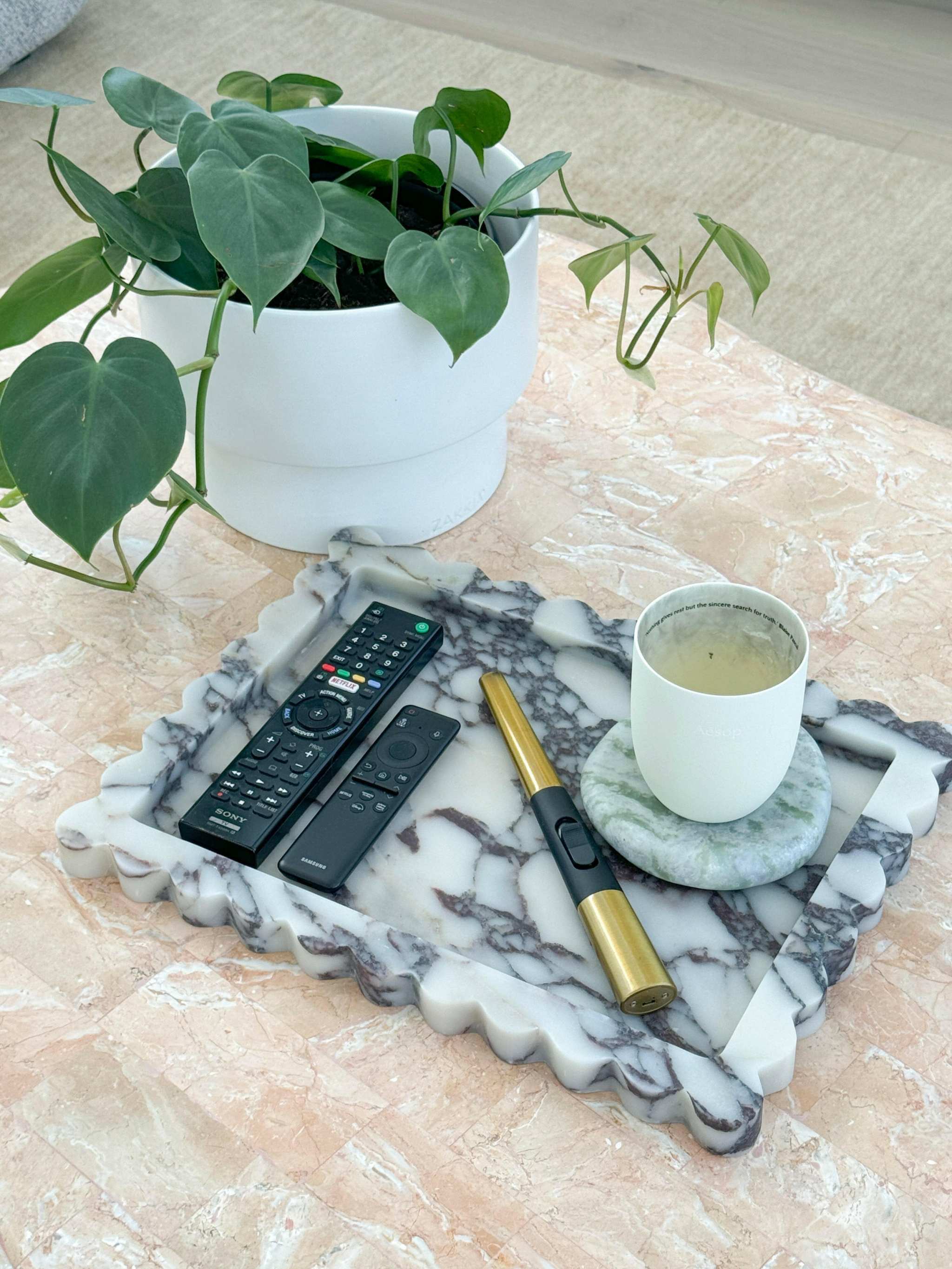 Large Palazzo scalloped marble bar tray in Viola marble with remote controls, lighter, and a candle on a coaster next to a potted green plant on a tiled surface