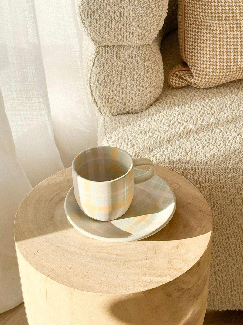 Robert Gordon hand-painted ceramic mug with pastel tartan design and matching saucer on wooden stool next to beige textured sofa
