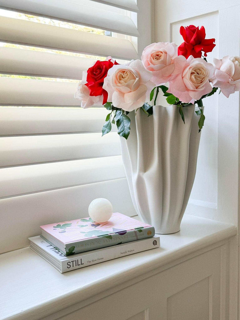 Small white onyx CoTheory Orbit Table Sculpture sphere displayed on stacked books next to a white vase with pink and red roses on a windowsill