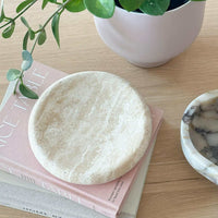 Small beige travertine tray with smooth curved edges on a pink book next to a white pot and plant, ideal decorative centrepiece tray