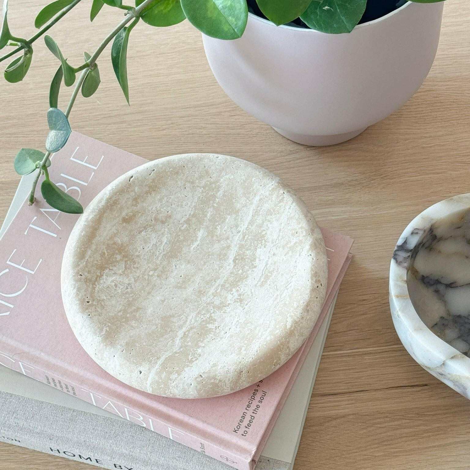 Small beige travertine tray with smooth curved edges on a pink book next to a white pot and plant, ideal decorative centrepiece tray
