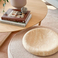 Large beige CoTheory Travertine sculpted tray with smooth curves on a textured round cushion next to a wooden coffee table with books and decor.