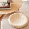 Large beige CoTheory Travertine Solstice sculpted tray with smooth curved design on textured round ottoman, next to wooden side table with books and decor.