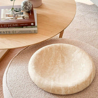 Large beige CoTheory Travertine Solstice sculpted tray with smooth curved design on textured round ottoman, next to wooden side table with books and decor.