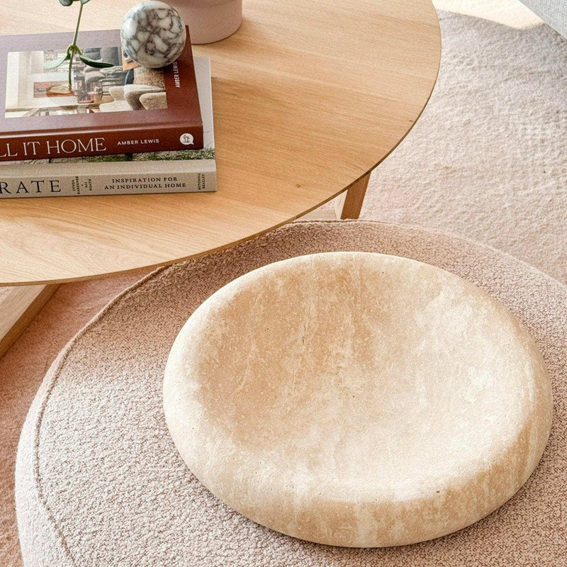 Large beige CoTheory Travertine Solstice sculpted tray with smooth curved design on textured round ottoman, next to wooden side table with books and decor.