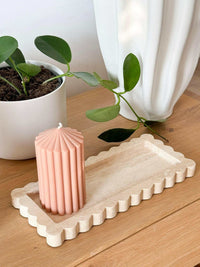 Beige travertine Palazzo Scalloped Marble Bar Tray with scalloped edges on wooden surface holding a pink ribbed candle, next to a green potted plant and white vase.