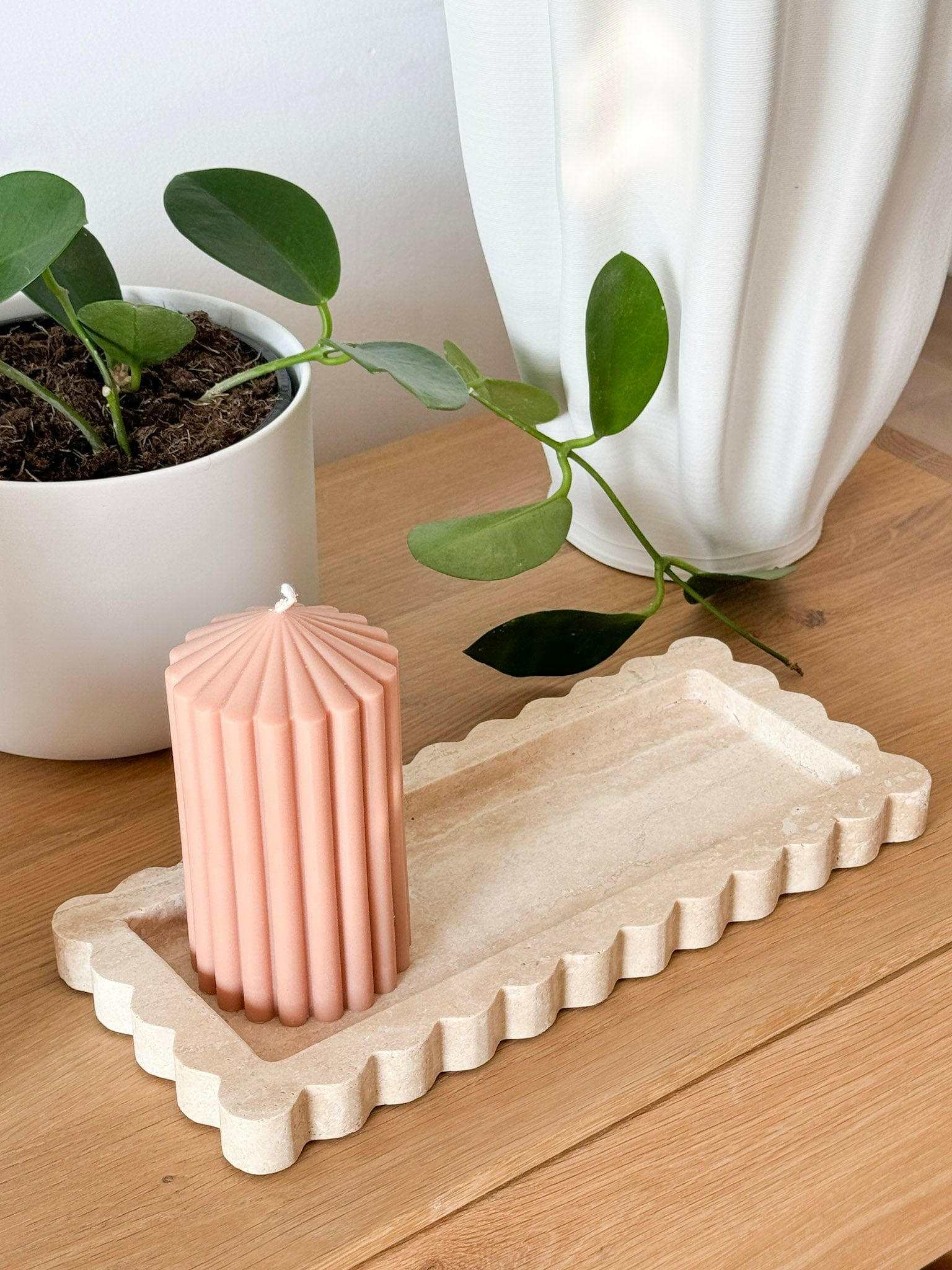 Beige travertine Palazzo Scalloped Marble Bar Tray with scalloped edges on wooden surface holding a pink ribbed candle, next to a green potted plant and white vase.