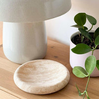 Small beige travertine tray with smooth curved design on wooden surface next to white lamp and green potted plant