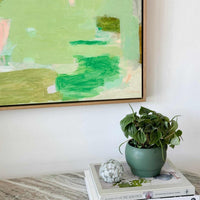 Limited edition abstract green and pastel canvas print "Lovely Day" by Vanni, framed in floating oak, displayed above a table with a green potted plant and books