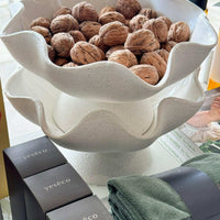 Ripple white ceramic pedestal bowl with textured exterior, holding walnuts, placed on a table with folded green towels and Yeseco branded boxes nearby
